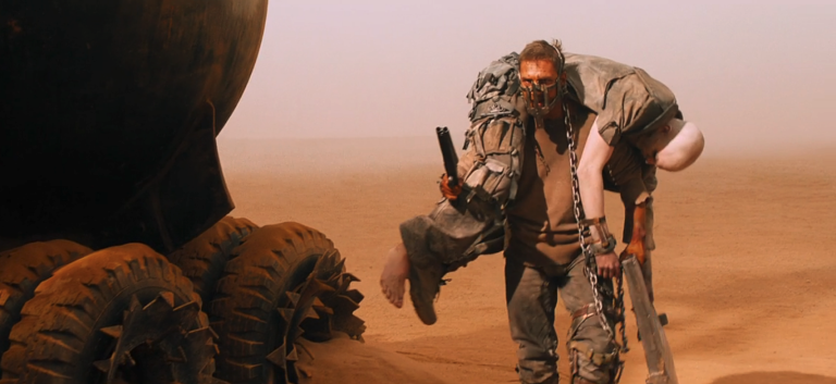 Mad Max Fury Road Max carries Nux and a shotgun toward Furiosa's war rig.