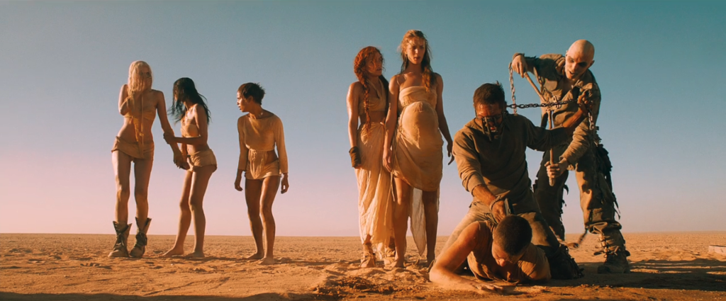 Max holds a gun to Furiosa's head while Nux uses bolt cutters to break the chain connecting him to Max.